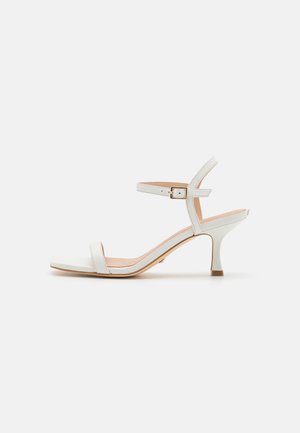 Hvid åben tå hælsandal til kvinder med ankelrem og lille guldbeklædt spænde, mellemhøj slank hæl, minimalistisk design.