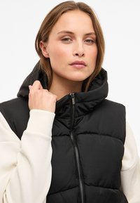 Juoda paminkštinta liemenė su aukšta apykakle ir užtrauktuko uždarymu. Medžiaga lygi, po apačia vilkimas baltas kontrastingas ilgomis rankovėmis megztinis.