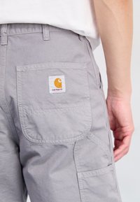 Pantalones cargo grises con un bolsillo trasero que presenta un parche de logo cuadrado en naranja y blanco. La textura de la tela parece duradera y suave.