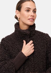 Veste en polaire texturée marron avec un col montant. Poignets en tricot côtelé. Matériau doux et duveteux avec un aspect laineux.