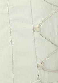 Mochila de tela de color claro con superficie texturizada, que cuenta con cordones elásticos y correas de webbing a lo largo del costado para ajuste o sujeción.
