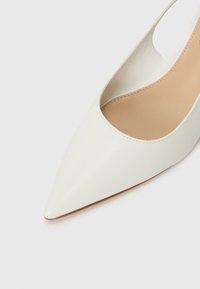 Valkoinen nahkainen kärkikengän malli, jossa on leikattu muotoilu sivuilla. Silkkiäinen pinta ja beige sisäpohja, jossa näkyy ompelusyksityiskohtia.