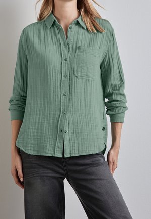 Kobieta ubrana w jasnozieloną koszulę o teksturze, z długim rękawem, zapinaną na guziki, z kieszenią na piersi oraz w ciemnoszare jeansy.