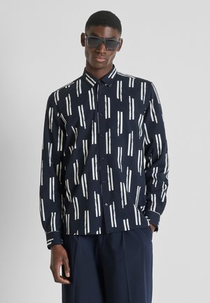 Uomo che indossa occhiali da sole neri e una camicia nera a maniche lunghe con bottoni e strisce verticali bianche a pennellate, in piedi davanti a uno sfondo grigio.