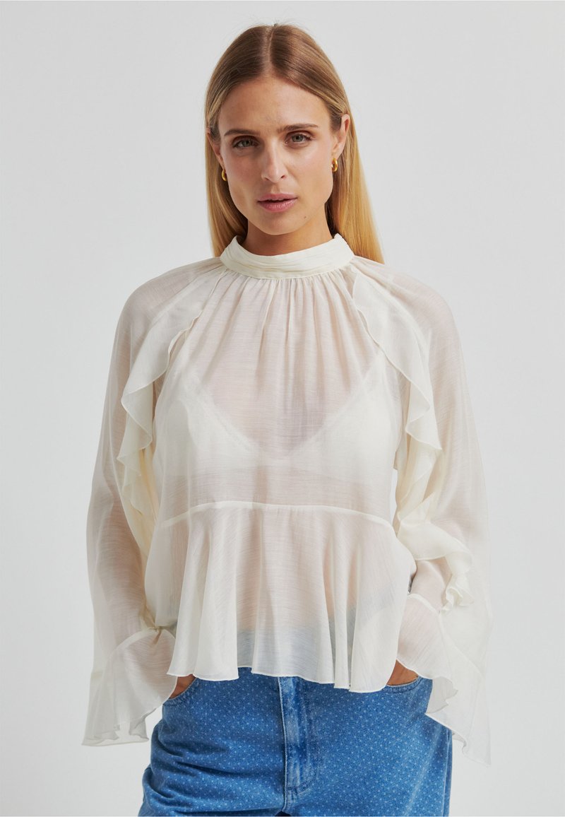 Blouse blanche semi-transparente avec manches à volants et col montant. Présente un ourlet évasé et un tissu texturé. Associée à un short en jean.
