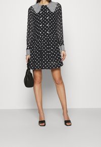 Robe noire à pois avec manches longues et col à carreaux. Comprend une jupe évasée, des détails à boutons et une finition texturée. Sac à main noir inclus.