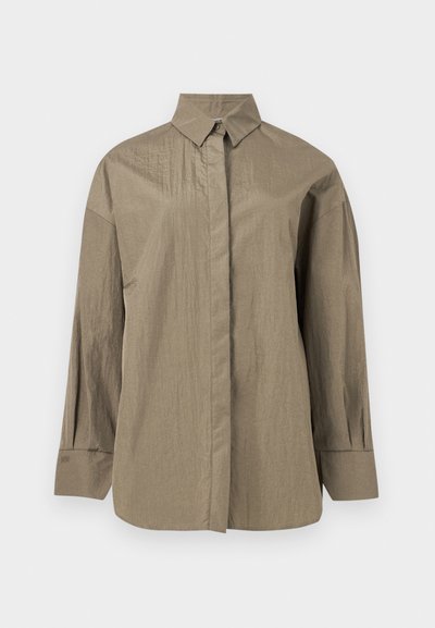 Túlméretezett, hosszú ujjú bézs ing hegyes gallérral, rejtett elülső gombokkal és összehúzott mandzsettával.