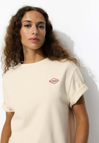 T-shirt beige à manches courtes avec un col rond et des manches roulées. Emblématique patch logo "Sunset" rouge et bleu sur la poche gauche. Texture douce.