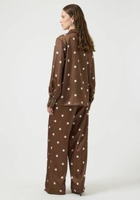 Brun langermet skjorte og matchende vid bukse, begge med kremfargede polkadotter. Jevn tekstur, lett glans, detaljer med knapper på mansjettene synlige.