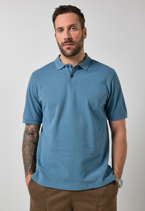 Uomo barbuto con braccio destro tatuato che indossa una polo blu e pantaloni marroni con le mani nelle tasche, su sfondo neutro.