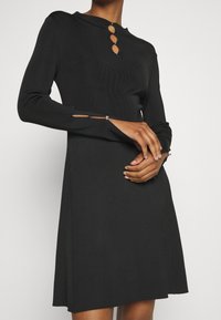 Robe noire à manches longues côtelées avec un col montant et trois boutons en forme de goutte au niveau du col. La robe présente des découpes latérales ornées de détails métalliques.