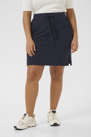 Žena v tmavomodrej sukni dĺžky po kolená s bočným rozparkom, bielom top-e a bielych masívnych teniskách, stojaca na jednoduchom svetlom pozadí.