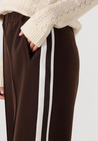 Bruine broek met een witte zijstreep, voorzien van een zijzak en een zachte textuur. Gepaard met een gebreide crème trui.