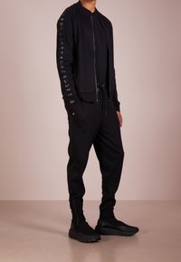 Completo sportivo nero composto da una giacca bomber con zip e strisce laterali a fantasia, abbinato a pantaloni jogger dalla vestibilità rilassata e sneakers nere.