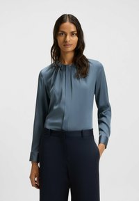 Lichtblauwe satijnen blouse met een geplooide hals, lange mouwen, voorkruisen en een soepele textuur, gecombineerd met donkere broek.