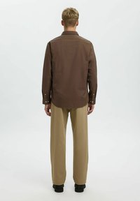 Camisa de algodón marrón con una textura sutil, puños con botón y dobladillo curvado, combinada con pantalones beige claros, vista desde la espalda.
