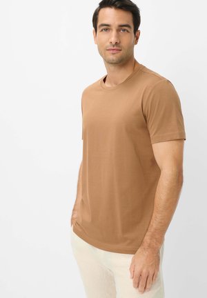 Junger Mann mit kurzen dunklen Haaren, der ein braunes T-Shirt mit kurzen Ärmeln und hellbeige Hosen trägt, steht vor einem schlichten weißen Hintergrund.