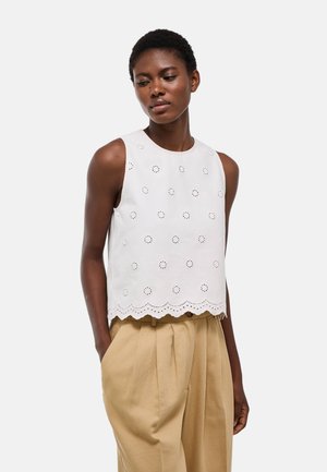 Femme portant un top blanc sans manches en broderie anglaise avec ourlet festonné et un pantalon plissé beige taille haute, mains dans les poches, sur fond uni.