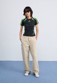 Schwarzes Sport-T-Shirt mit grünen Akzenten, kombiniert mit beigen weiten Hosen. Das Model trägt elegante schwarze Sonnenbrillen und weiße Sneakers mit schwarzen Streifen.