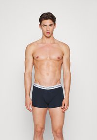 Boxers en coton bleu foncé avec une ceinture blanche portant le logo "TOMMY HILFIGER". Modèle debout, mettant en valeur l'ajustement et le design.