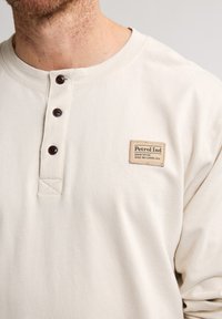 Lichtbeige henley-shirt met lange mouwen, voorzien van drie donkerbruine knopen en een rechthoekig geweven label met de tekst "Petrol Ind". Soepele textuur.