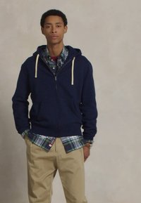 Marine blouson met rits en crème kleurige koordjes, gelaagd over een groen en blauw geruit shirt, gecombineerd met beige broek en een polsaccessoire.