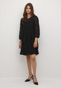 Schwarzes Spitzenkleid mit Dreiviertelärmeln, einem runden Ausschnitt und einem weit auslaufenden Saum. Verfügt über aufwendige Blumenmuster und eine leichte Textur.
