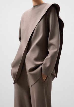 Personne portant un top oversize taupe avec un rabat asymétrique sur une épaule, assorti à un pantalon large coordonné, mains dans les poches.