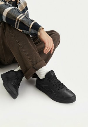 Person wearing brown pants, white socks, and black Reebok sneakers, sitting cross-legged on a light background.