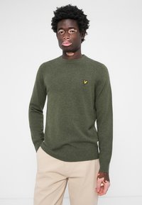 Pull vert olive en tissu doux, avec des manches longues et un col ras du cou. Présente un petit logo jaune sur la poitrine gauche.