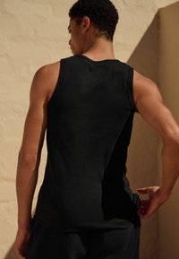 Schwarzes geripptes Tanktop mit taillierter Passform, das Details im Rücken- und Schulterbereich zeigt, kombiniert mit strukturierten, dunklen Shorts. Heller Hintergrund.