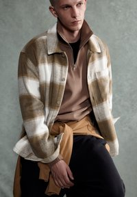 Ung man klädd i en beige och vit rutiga jacka över en brun dragkedjetröja, med en tan sweatshirt knuten runt midjan och svarta byxor.