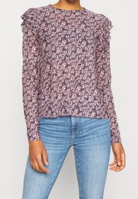 Langärmlige Bluse in Dunkelpurpur mit floralem Muster in Pink und Weiß, mit Rüschenschulterpartien und rundem Ausschnitt.