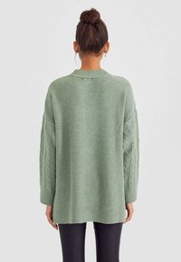 Oversized groene gebreide trui met kabelgebreide mouwen, geribbeld zoom en ronde hals. Zachte textuur met een relaxte pasvorm, geschikt voor lagen.
