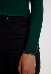Green ribbed long-sleeve top with scalloped hem, paired with black jeans. Focus on textured fabric and slim fit.