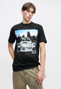 Melna kokvilnas T-kreklu ar krāsainu grafikā attēlotu boomboksu un pelēko suņu siluetu uz pilsētas ainavas fona.