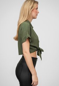 Cămașă scurtă de culoare verde măsliniu cu mâneci scurte și un nod în față, asortată cu pantaloni negri cu talie înaltă, având o textură netedă și un fermoar.