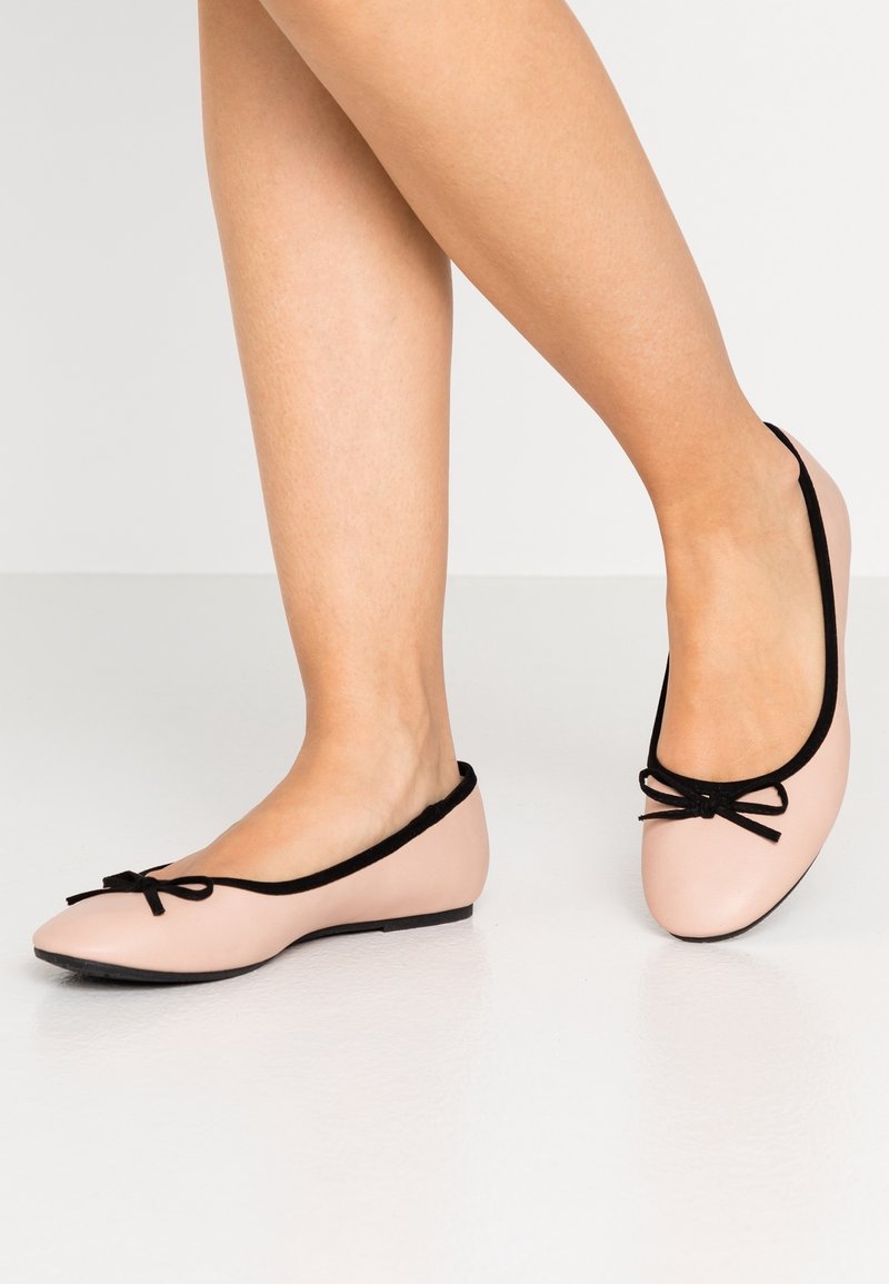 Ballerines roses pâles avec un liseré noir et un détail nœud, fabriquées en matériau lisse. Le design présente un profil bas et un bout arrondi.