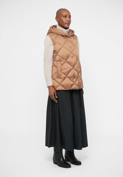 Gilet matelassé tan avec capuche, associé à un pull crème et une jupe plissée noire. Des bottines noires viennent compléter la tenue.