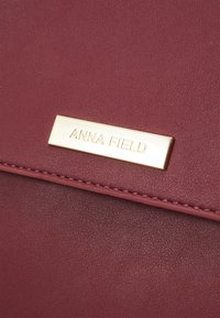 Kastanjebrun faux lædertaske med et guldtonet metalbadge indgraveret med "ANNA FIELD" og subtile stikninger langs kanten.