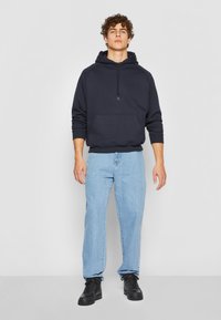 Felpa con cappuccio navy e tasca frontale, abbinata a jeans azzurri chiari e sneakers nere. Il tessuto sembra morbido con una vestibilità comoda.