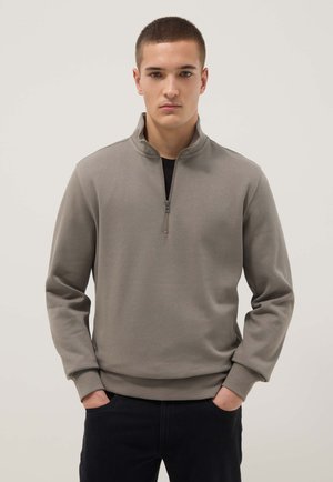 Jeune homme aux cheveux courts portant un pull à fermeture éclair taupe et un pantalon noir, debout les mains dans les poches devant un fond uni.