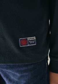 Mörkblå sweatshirt i texturerat tyg med en röd och vit märkeslapp som visar "Superdry Vintage Athletic Outfitters".