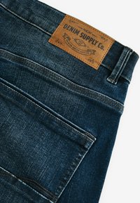 Donkerblauwe denim jeans met een leren merklabel met de tekst "DENIM SUPPLY Co." en zichtbare stiksels op de tailleband en zakken.