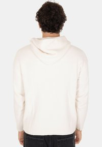 Felpa bianca a maglia con finitura testurizzata, dotata di cappuccio con cordino e polsini a coste, indossata con pantaloni neri. Vista posteriore.