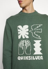 Homme portant un pull en maille verte avec des formes abstraites blanches et le texte "QUIKSILVER" sur le devant, cadré au niveau du cou et de la taille.