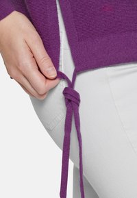 Lila stickad tröja med en texturerad finish, med en knytbar dragsko vid fållen, bärs över vita byxor. Närbild på en hand som justerar knuten.