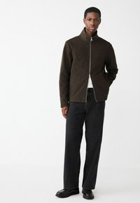 Tmavohnedá bunda na zips s vysokým golierom, nosená cez bielu košeľu a kombinovaná s čiernymi prúžkovanými nohavicami a čiernymi topánkami.