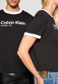 Juodos medvilnės marškiniai su baltais akcentais, ant kurių matyti "Calvin Klein endless city" logotipas. Viena maikutė įdėta į džinsinius šortus.