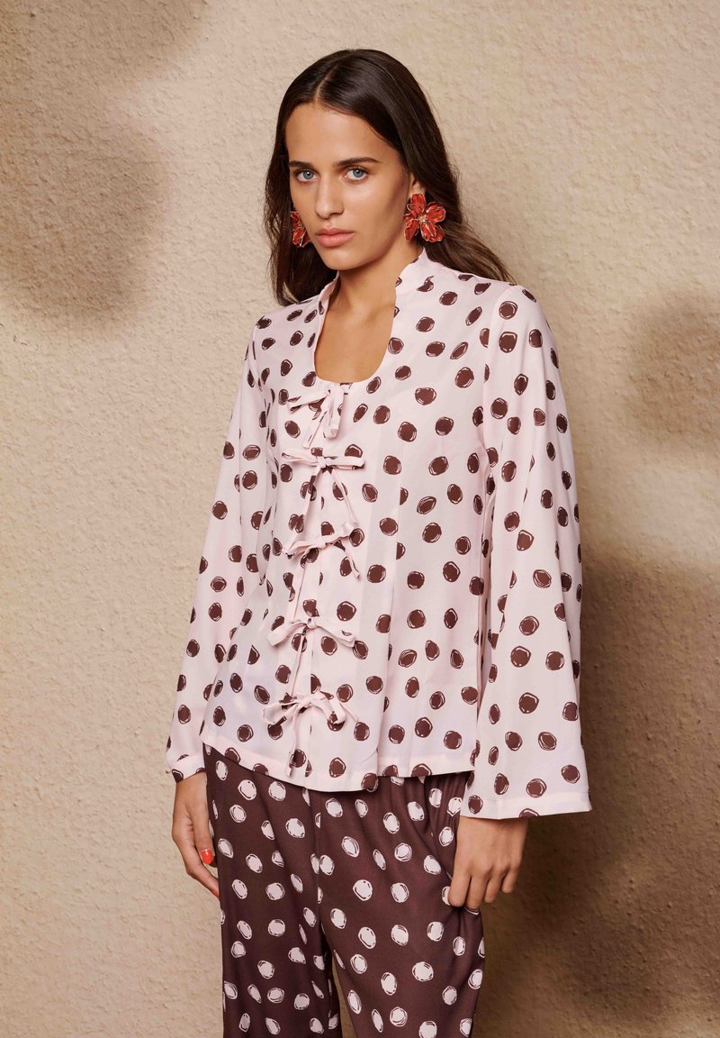 Blouse rose clair à pois marron foncé, avec un panneau à volants et un décolleté ouvert. Associée à un pantalon assorti à pois.